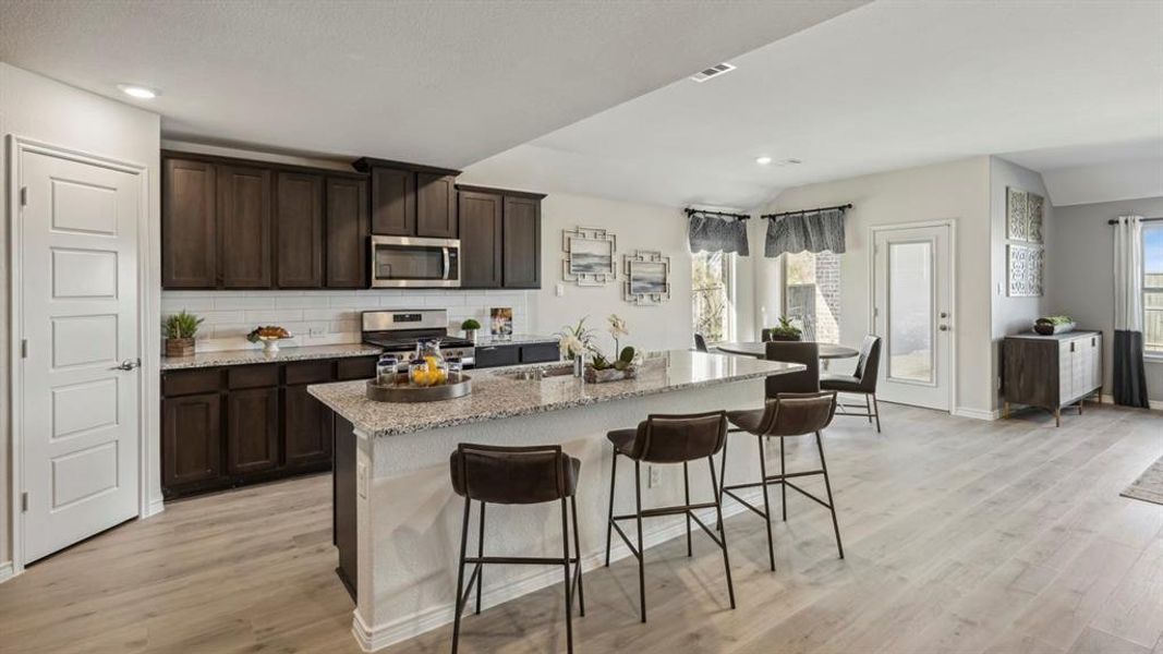This open-concept kitchen features dark wood cabinetry, stainless steel appliances, a white subway tile backsplash, and light granite countertops, including a spacious island with seating