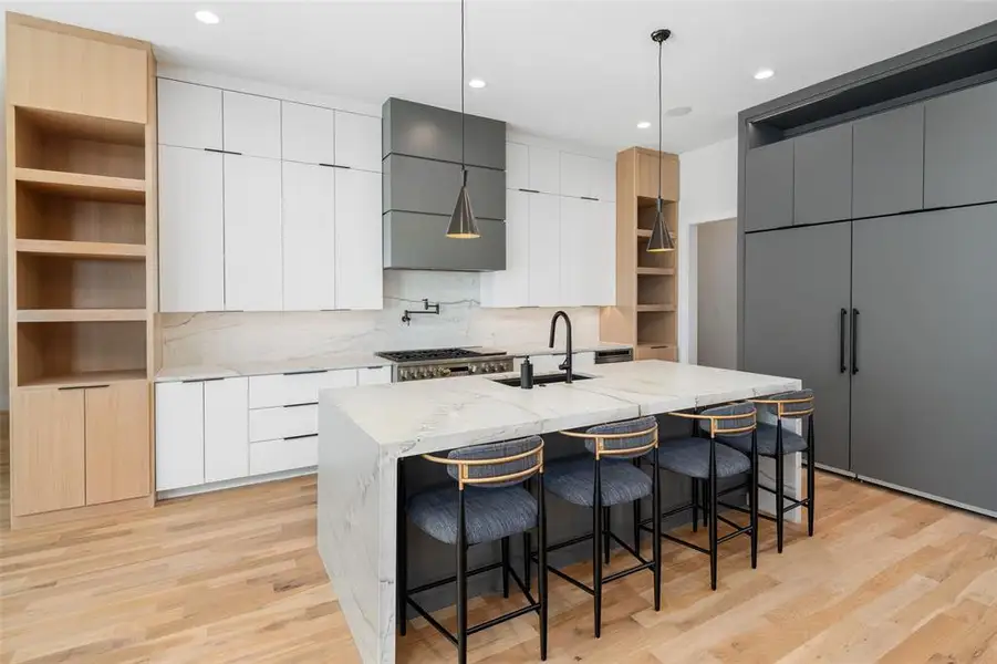 Designer kitchen featuring custom cabinetry with clean, modern lines, warm wood accents, and sleek hardware. A large waterfall island with seating anchors the space, complemented by Taj Mahal quartzite countertops, a commercial vent hood, and high-end appliances, creating a perfect blend of style and functionality.