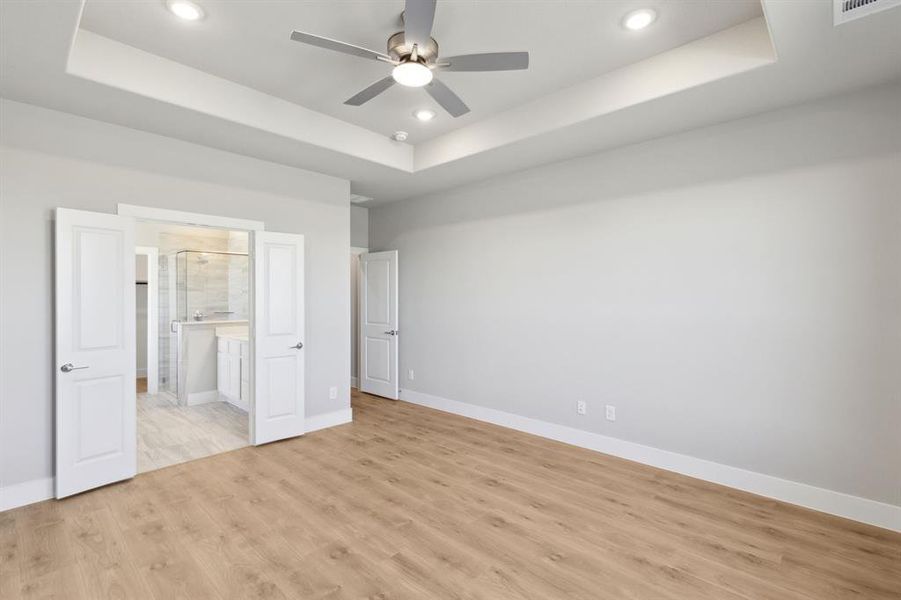 Unfurnished bedroom with ensuite bath, ceiling fan, light hardwood / wood-style flooring, and a tray ceiling