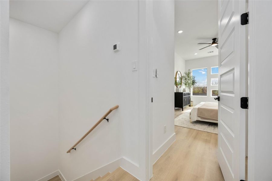 A bright upper-level landing features clean white walls and warm wood-look flooring, opening into a spacious bedroom with large windows, contemporary finishes, and abundant natural light for a welcoming transition between spaces.