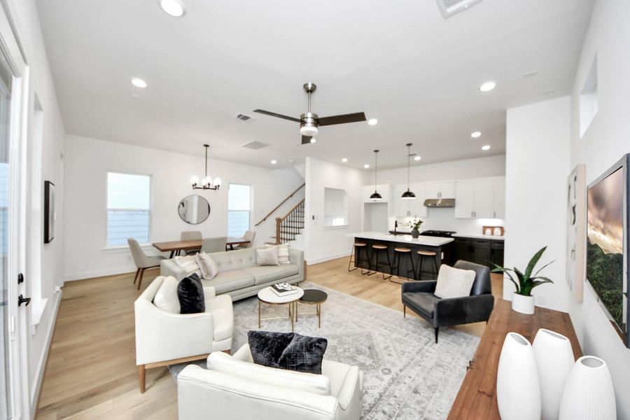 This photo showcases a modern, open-concept living space featuring a bright living room with a ceiling fan, a dining area, and a sleek kitchen. The kitchen has a large island with barstools, pendant lighting, and white cabinetry. The room is adorned with stylish furniture, wood flooring, and ample natural light. This photo showcases a modern, open-concept living space featuring a bright living room with a ceiling fan, a dining area, and a sleek kitchen. The kitchen has a large island with barstools, pendant lighting, and white cabinetry. The room is adorned with stylish furniture, wood flooring, and ample natural light.