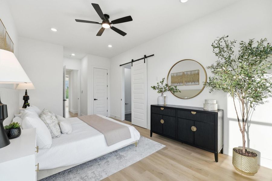 Spacious primary bedroom featuring wood-look flooring, recessed lighting, a modern ceiling fan, and a stylish barn-door entry, complemented by clean lines and abundant natural light for a serene, contemporary retreat.