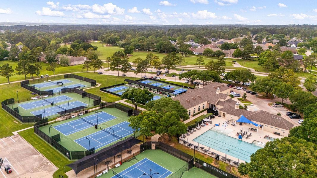 This photo showcases a vibrant community setting featuring multiple tennis courts, a large swimming pool, and a clubhouse. The area is surrounded by lush greenery and residential homes, offering recreational amenities in a serene neighborhood environment.
