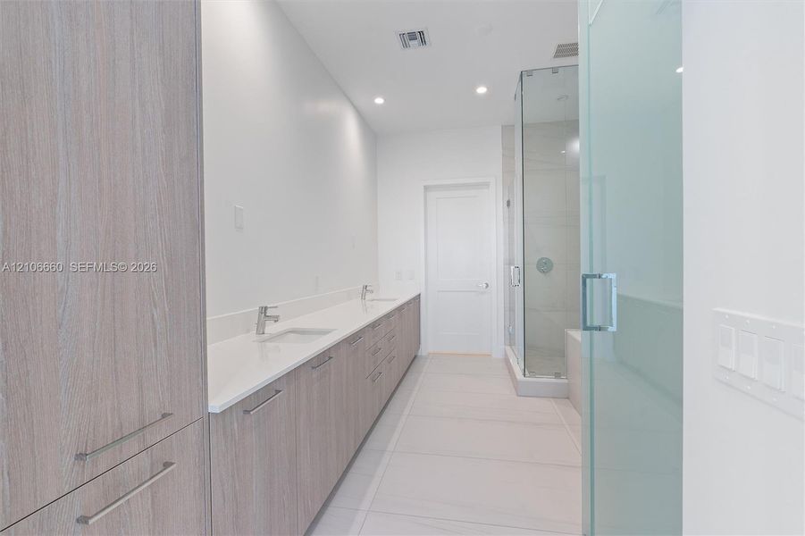Dual-sink vanity with wood-finish cabinetry, light stone countertop, walk-in glass-enclosed shower, and tile flooring
