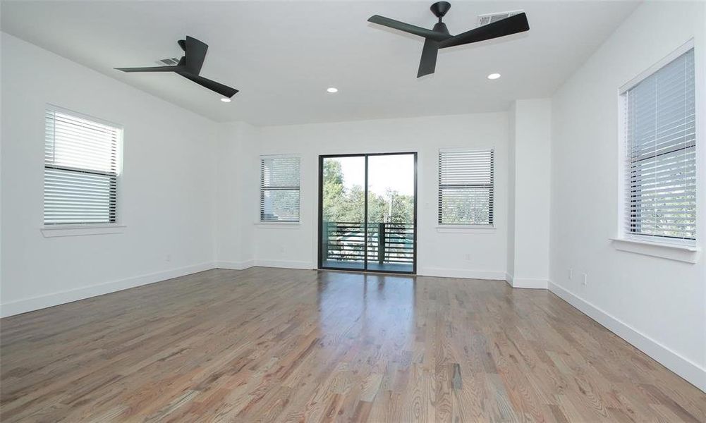 This spacious, bright room features hardwood flooring, large windows, and a glass door leading to a second story walk out balcony. It includes modern ceiling fans and recessed lighting, perfect for a comfortable and airy living space.