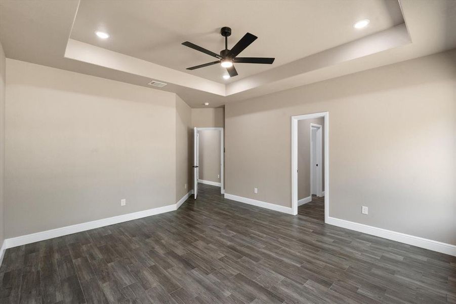 Primary bedroom featuring tray ceiling with recessed lighting and a ceiling fan, wood-finish flooring, two windows, and neutral wall tones. Individual bedroom remote for integrated lighting.