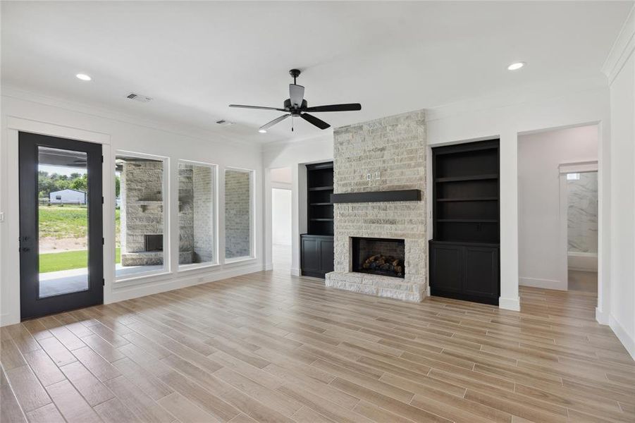 Unfurnished living room with light wood-type flooring, crown molding, built in features, ceiling fan, and a stone fireplace