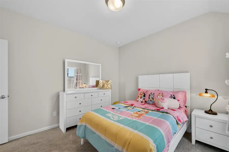 This cozy bedroom features a playful theme with colorful bedding and plush toys. It's currently adorned with a white dresser with a mirror and a matching nightstand, all set against neutral walls and carpet, creating a warm and inviting space. This cozy bedroom features a playful theme with colorful bedding and plush toys. It's currently adorned with a white dresser with a mirror and a matching nightstand, all set against neutral walls and carpet, creating a warm and inviting space.