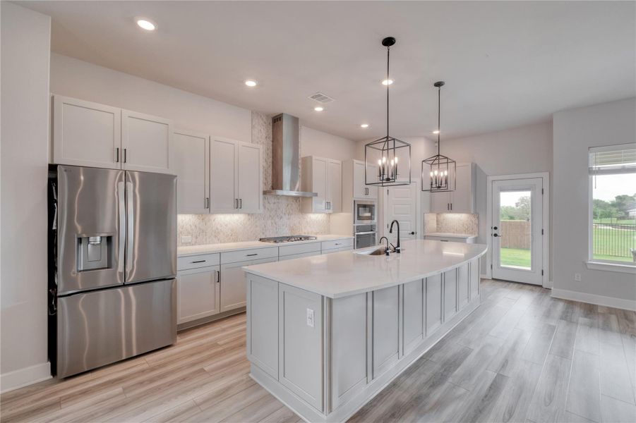 Spacious kitchen featuring an expansive central island with a built-in sink, stainless steel appliances, light wood-finish flooring, white cabinetry, and recessed lighting