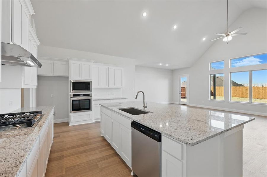Kitchen featuring white cabinets, light wood-style floors, stainless steel appliances, light stone counters, and an island with sink