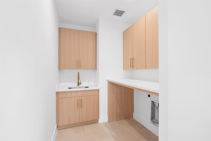 Well-appointed utility area featuring light wood cabinetry with gold-toned hardware, a white countertop, and a utility sink with a gold-toned faucet