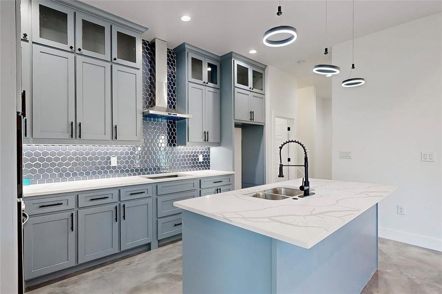 Kitchen featuring wall chimney range hood, hanging light fixtures, gas stovetop, light stone countertops, and a kitchen island with sink