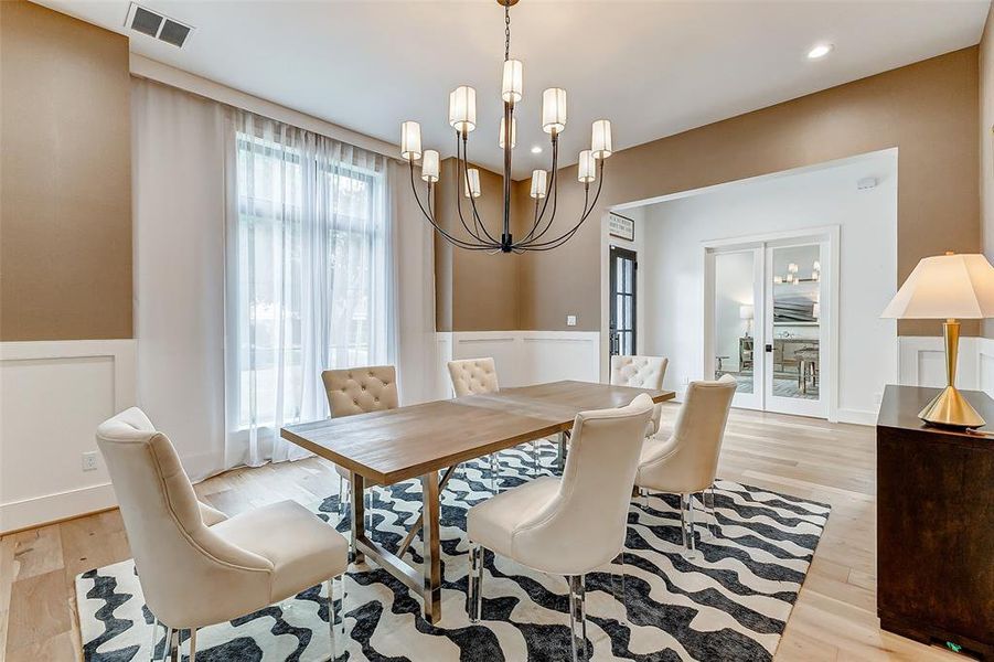 This dining room features elegant lighting, a modern wooden table with six upholstered chairs, and large windows with sheer curtains. The space is accented with a stylish rug and neutral tones, creating a warm, inviting atmosphere.