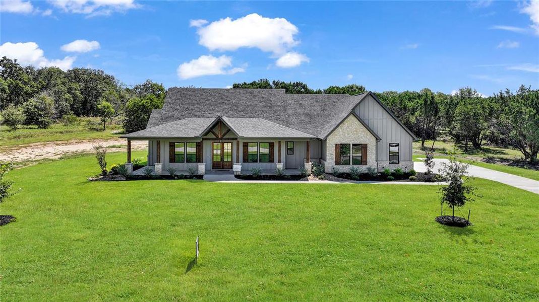 Modern farmhouse with covered porch, a front yard, roof with shingles, and stone siding