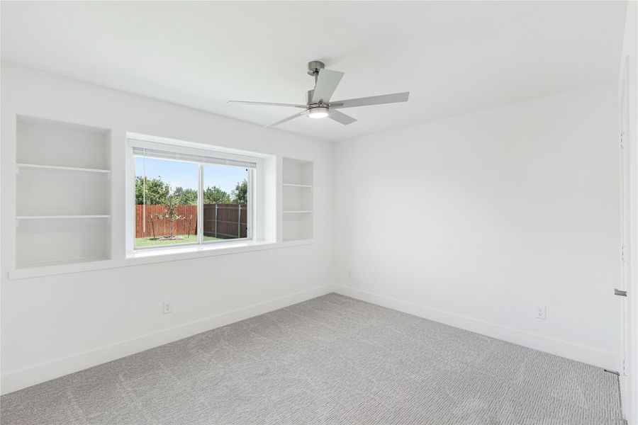 Empty room with gray carpet, ceiling fan, built-in shelves, and a large window overlooking a fenced backyard