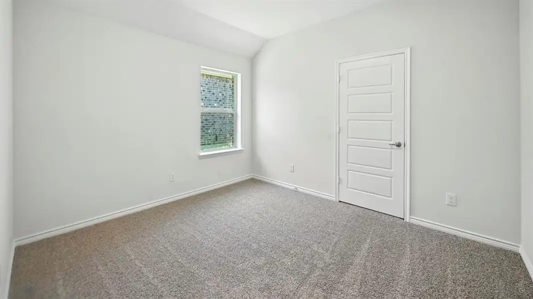 Spacious room featuring neutral carpeting, white walls, a single window with exterior brick scenery, a white paneled door with a brushed nickel handle, and white baseboards Spacious room featuring neutral carpeting, white walls, a single window with exterior brick scenery, a white paneled door with a brushed nickel handle, and white baseboards
