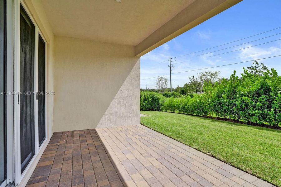 Covered patio with paver flooring, stucco exterior, sliding glass doors, and a manicured lawn with privacy hedges