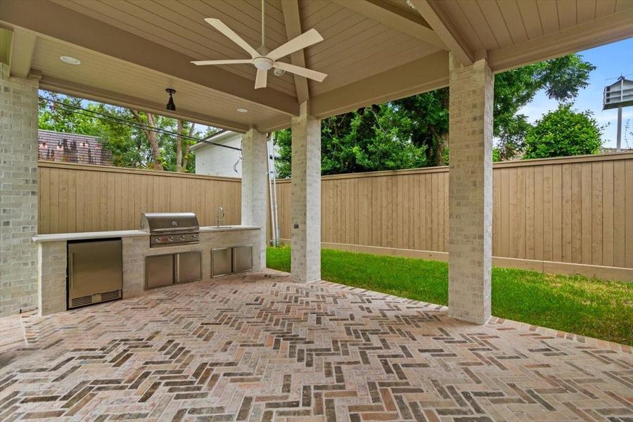Outdoor kitchen features herringbone brick floors, refrigerator, built in gas grill, sink, fan
