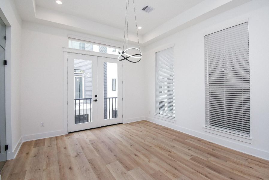 Bright room with modern light fixture, light wood flooring, and large windows featuring white blinds. French doors open to a small balcony, enhancing natural light. Ideal for dining or living space.