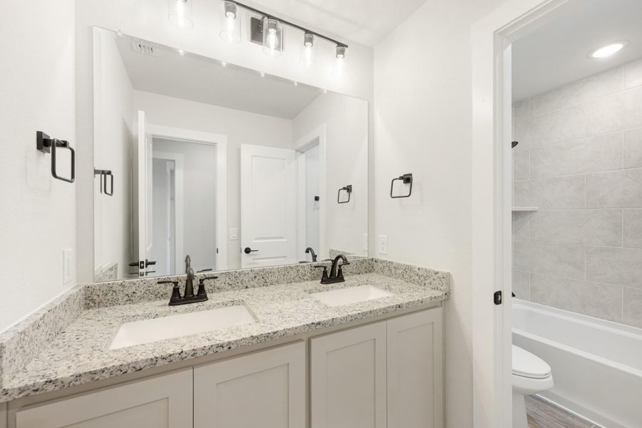 Bathroom with double sink vanity, granite countertop, large mirror, and tiled shower-tub combo visible through doorway.