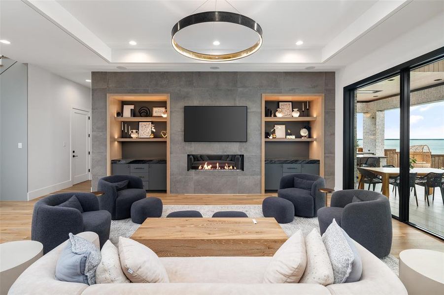 Beautifully designed living room centered around a sleek linear fireplace set within a dramatic floor-to-ceiling tile feature wall. Custom built-in cabinetry and shelving with accent lighting provide both style and function, while the modern chandelier and real hardwood floors complete the warm yet contemporary space.