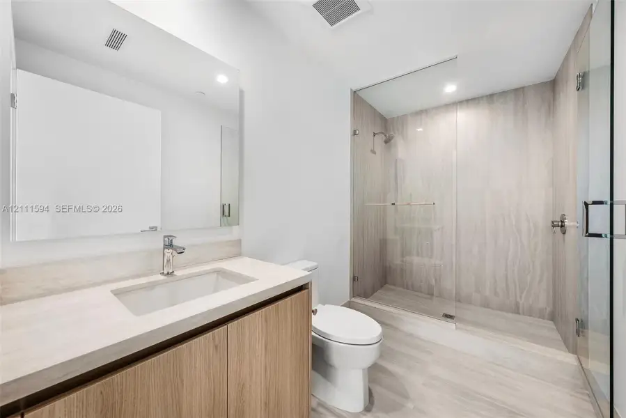 Bathroom featuring a wood-finish vanity with an integrated countertop, a frameless glass-enclosed walk-in shower with floor-to-ceiling stone-style tile, and a wall-mounted mirror Bathroom featuring a wood-finish vanity with an integrated countertop, a frameless glass-enclosed walk-in shower with floor-to-ceiling stone-style tile, and a wall-mounted mirror