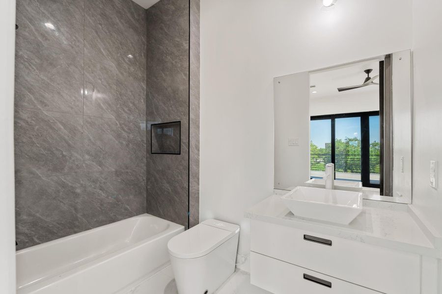 This secondary bath features large-format gray tile running floor-to-ceiling on the tub surround wall, a  tub and shower combination with refined fixtures, and a floating white vanity with a vessel sink.