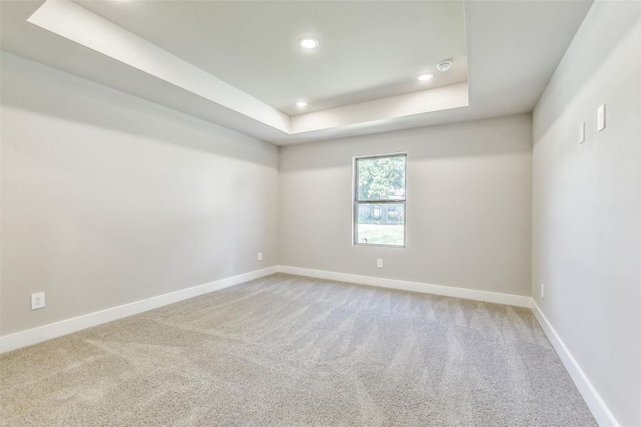 Spacious primary bedroom with tray ceiling detail, soft carpet flooring and a window overlooking the front yard.