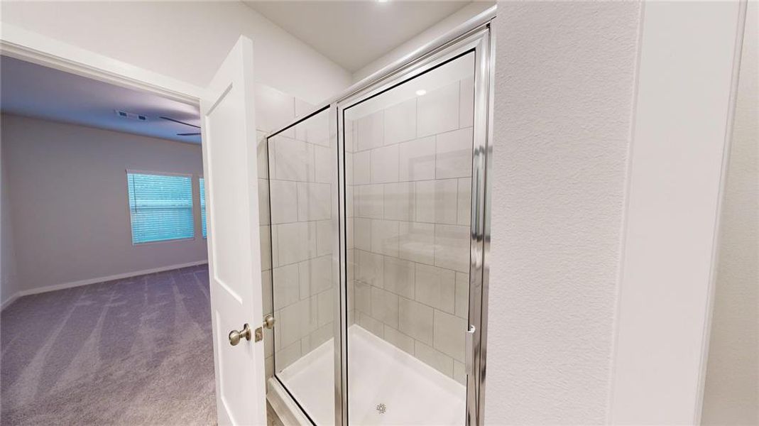 This photo shows a modern bathroom with a glass-enclosed shower featuring sleek tile work. It connects to a carpeted bedroom with a large window, offering a bright and inviting layout.