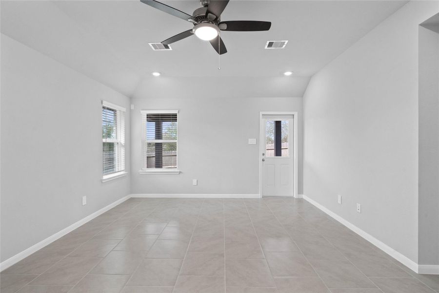 A bright and airy open-concept space featuring tile flooring, sloped ceilings with recessed lighting and ceiling fan, abundant natural light from large windows (fitted with blinds), and an access door to the back covered patio.