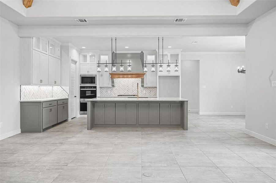 Kitchen with backsplash, white cabinets, custom exhaust hood, gray cabinetry, and a center island with sink Kitchen with backsplash, white cabinets, custom exhaust hood, gray cabinetry, and a center island with sink