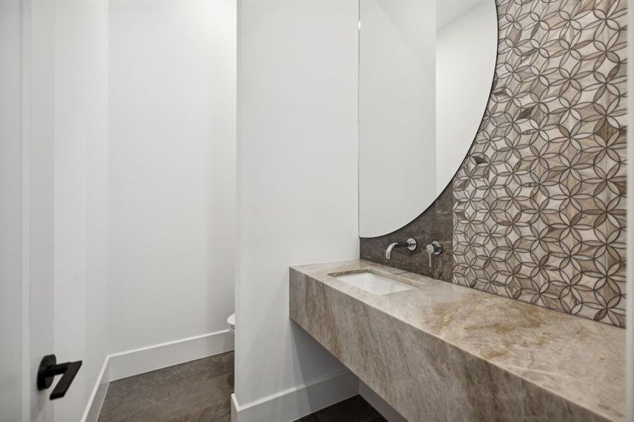 The floating stone vanity with a rectangular under-mount sink dominates the space. The stone hasa warm, neutral tone with subtle veining, while the modern faucet with a single lever handle is mounted onthe geometric pattern wall covering.