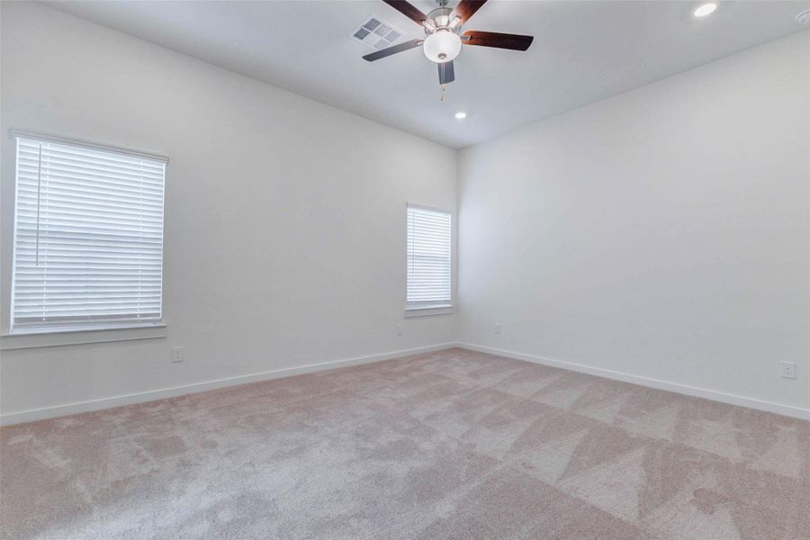 Spacious primary bedroom with soft carpet, high ceilings, ceiling fan, recessed lighting, and two windows bringing in natural light. The neutral walls and generous floor space create a calm, comfortable retreat with plenty of room for a full bedroom setup, sitting area, or personalized décor.