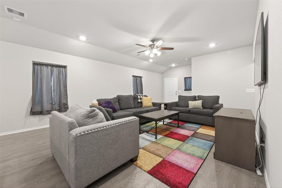 This inviting living room features modern gray sofas, a colorful area rug, and a ceiling fan, creating a cozy atmosphere. It's well-lit with recessed lighting and has a mounted TV for entertainment.