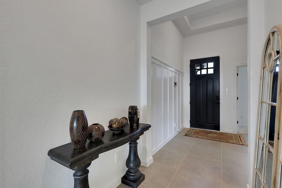 This entryway features a stylish black front door, light tile flooring, and a decorative console table with vases. The space is bright with white walls and wainscoting, creating a welcoming and modern look.