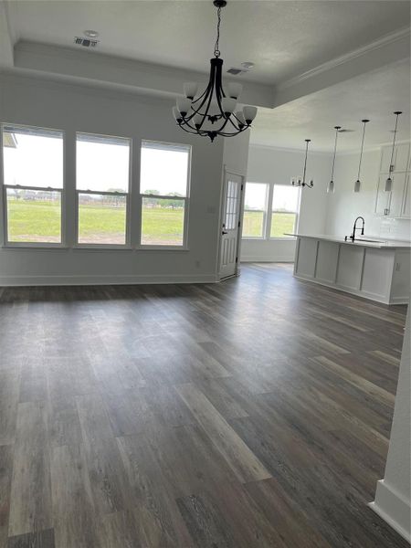 This photo showcases a bright, open-concept living space with large windows, medium gray/wood water proof plank flooring, and modern light fixtures. It features a spacious kitchen area with a white island and pendant lighting, offering a view of a green outdoor space.
