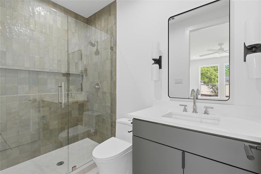 Bathroom featuring a glass-enclosed shower with tile surround, a rectangular vanity mirror, dual wall-mounted sconces, a contemporary toilet, and a vanity with a white countertop and gray cabinetry