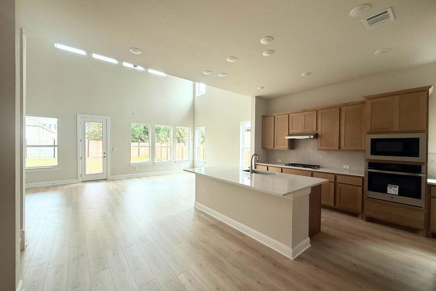 Open-concept kitchen and living area featuring high ceilings, wood-finish flooring, a kitchen island with sink, and stainless steel appliances including a built-in oven and microwave Open-concept kitchen and living area featuring high ceilings, wood-finish flooring, a kitchen island with sink, and stainless steel appliances including a built-in oven and microwave