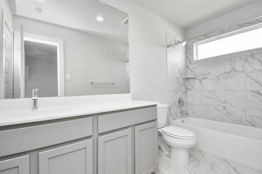 The secondary bath boasts stylish tile flooring, a bath/shower combo with a sleek tile surround, light wood cabinets, and a stunning light countertop. Sample photo of completed home with similar floor plan. As-built interior colors and selections may vary.