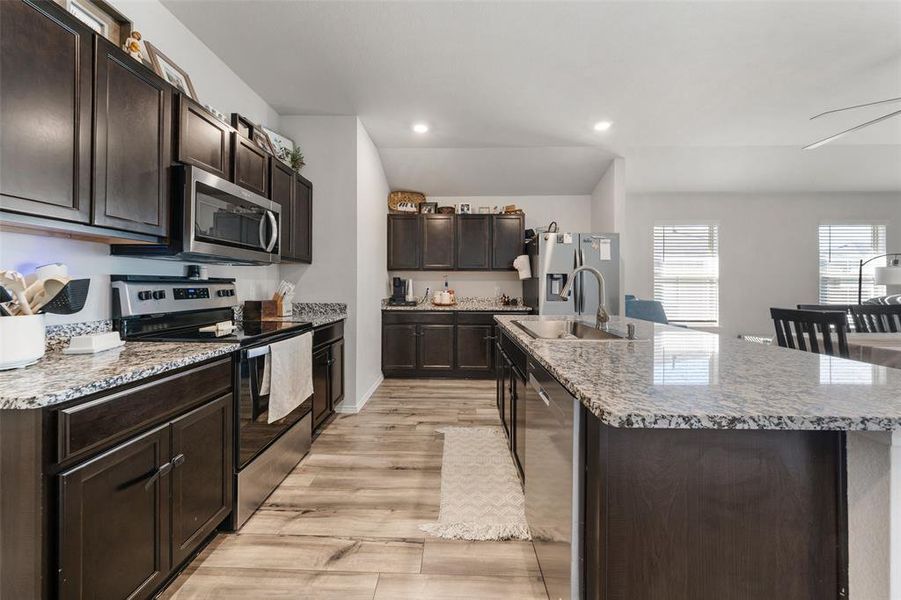Kitchen with dark brown cabinetry, appliances with stainless steel finishes, an island with sink, light wood-style floors, and light stone countertops