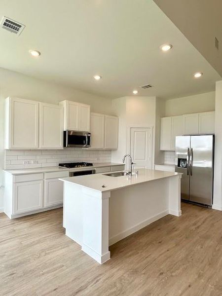Modern kitchen featuring a central island with a sink, white cabinetry, light-toned countertops, a subway tile backsplash, and wood-finish flooring