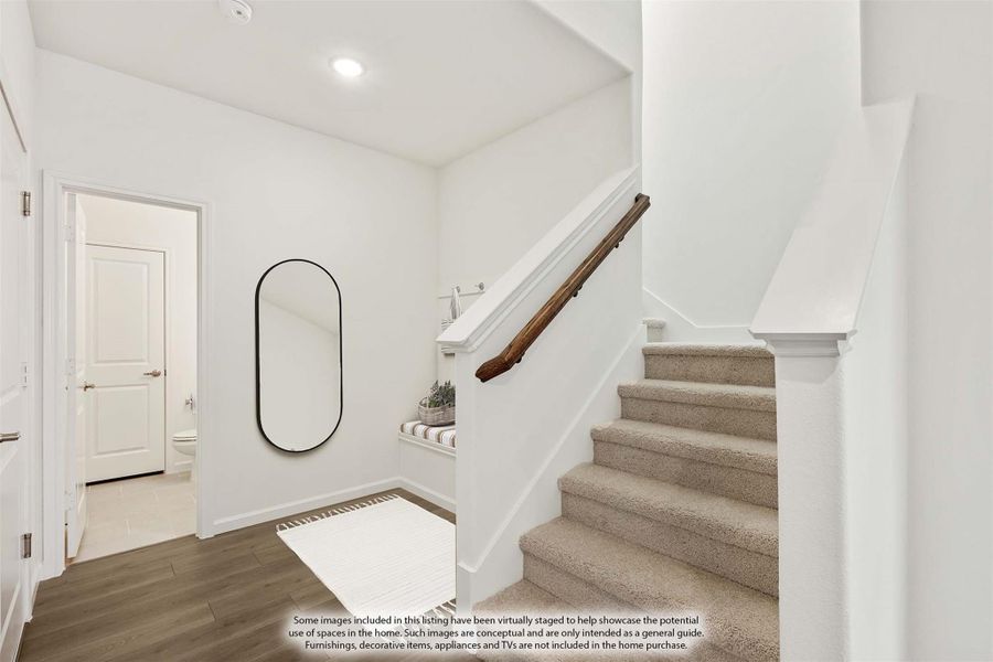 Entryway hallway with carpeted staircase, wood handrail, oval mirror, and hardwood floors leading to a bathroom