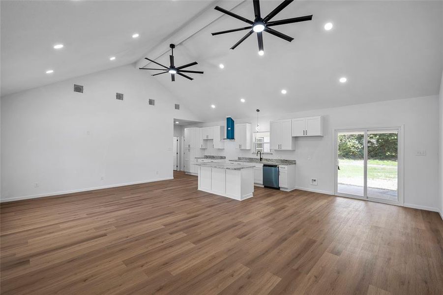 Open-concept living space featuring vaulted ceilings with recessed lighting, wood-finish flooring, a kitchen island with stone countertops, white shaker cabinetry, and sliding glass doors to the exterior Open-concept living space featuring vaulted ceilings with recessed lighting, wood-finish flooring, a kitchen island with stone countertops, white shaker cabinetry, and sliding glass doors to the exterior