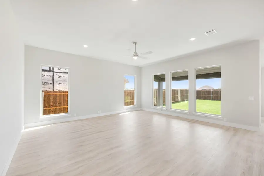 Empty living room with light wood floors, ceiling fan, and large windows overlooking a fenced backyard