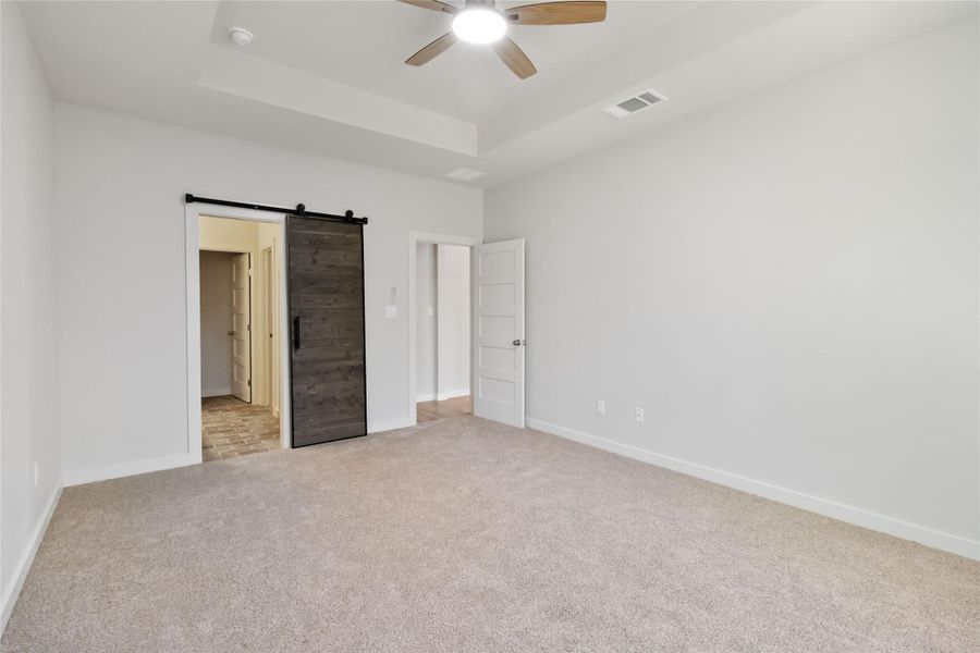 Unfurnished bedroom with light colored carpet, a tray ceiling, a barn door, and a ceiling fan