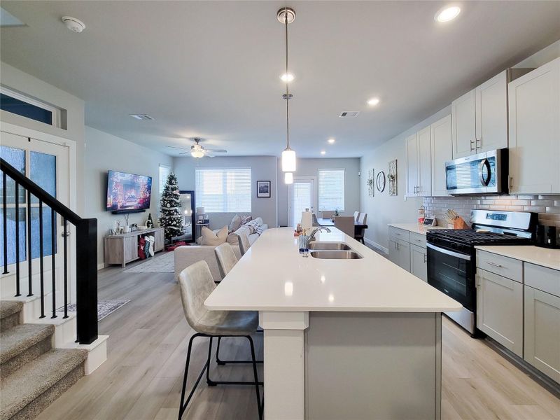 Open-concept living area with a kitchen featuring a large island, stainless steel appliances, and white cabinetry. The space is bright with ample natural light and includes a cozy living room, dining area, and a staircase leading upstairs.