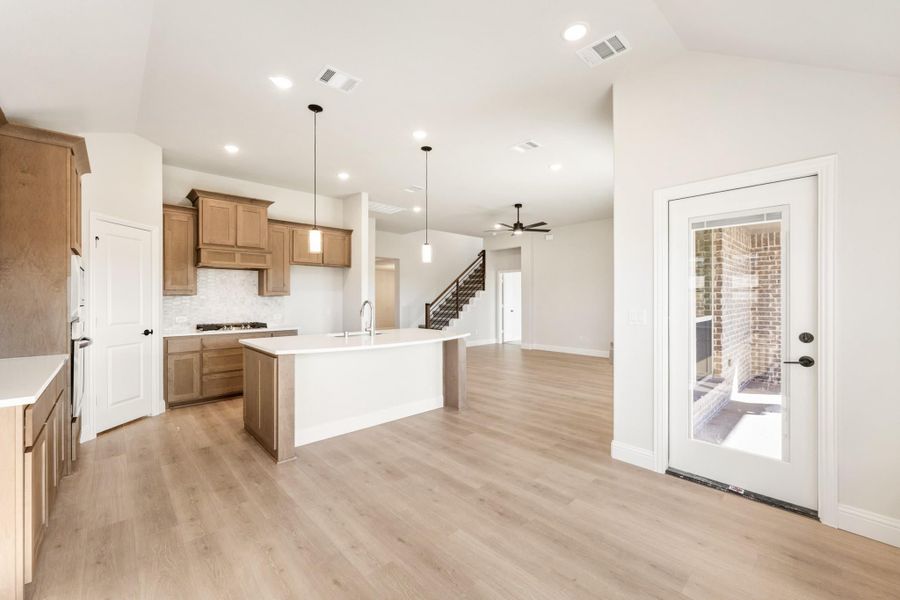 Open kitchen with wood cabinets, center island, pendant lights, and light hardwood floors open to living area with staircase.