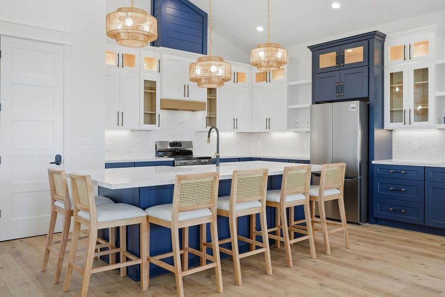 This kitchen features a modern design with navy and white cabinetry, a large island with seating, and stainless steel appliances. Elegant pendant lights and wood flooring add warmth and style.