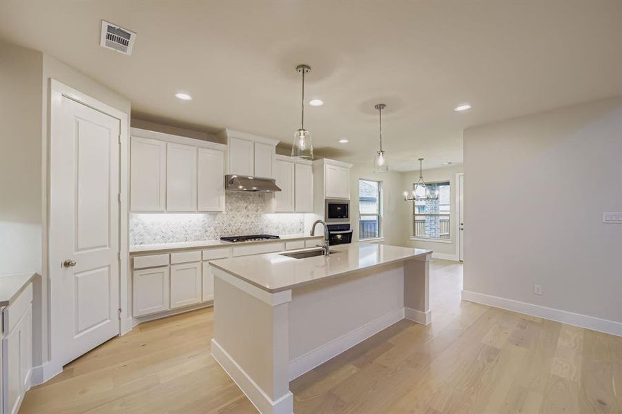 Kitchen featuring decorative backsplash, a center island with sink, decorative light fixtures, white cabinets, and light wood finished floors Kitchen featuring decorative backsplash, a center island with sink, decorative light fixtures, white cabinets, and light wood finished floors