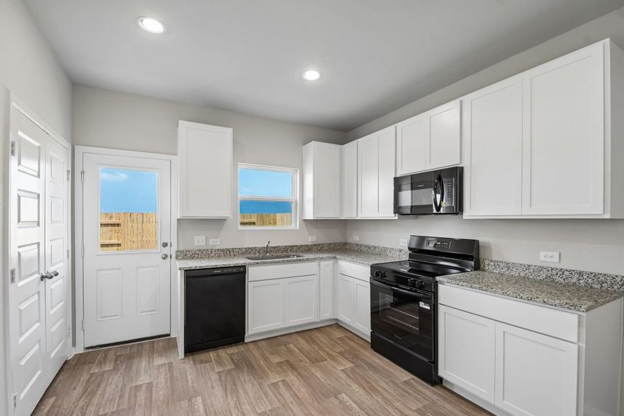 Image of home L-shaped kitchen with door to the backyard, window over kitchen sink overlooking the backyard. White cabinets, pantry doors, and walls with wood-look flooring. Granite countertops and black dishwasher, stove, and microwave.